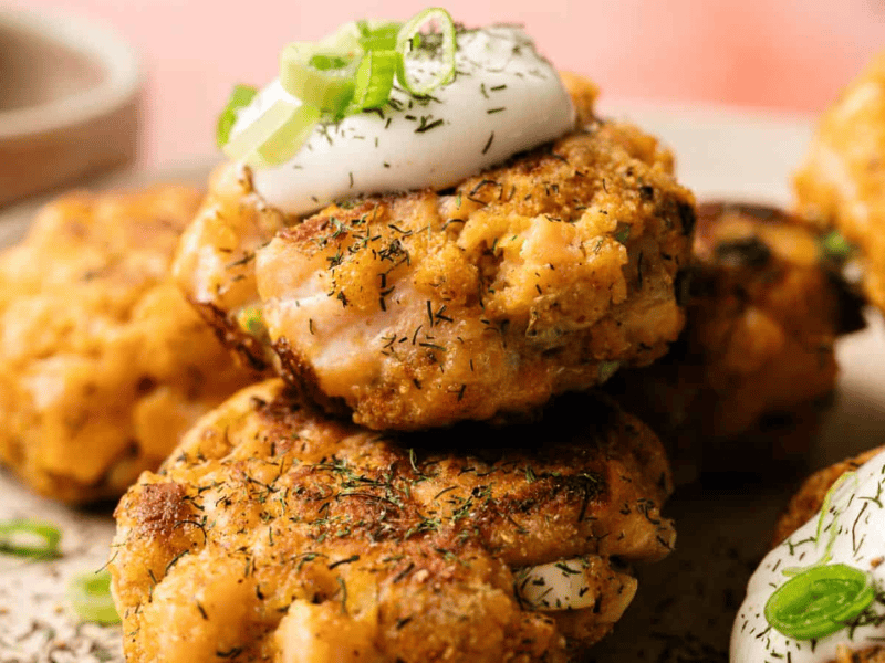 Close-up of golden-brown salmon patties topped with sour cream and chopped green onions, garnished with dried herbs on a plate.