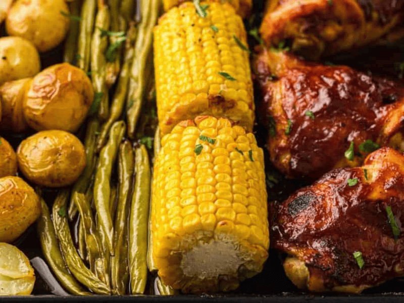 Roasted baby potatoes, green beans, corn on the cob, and barbecue chicken arranged on a sheet pan.