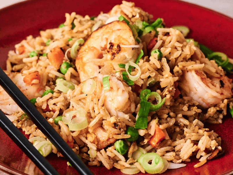 A red plate of shrimp fried rice garnished with chopped green onions, with black chopsticks resting on the side.