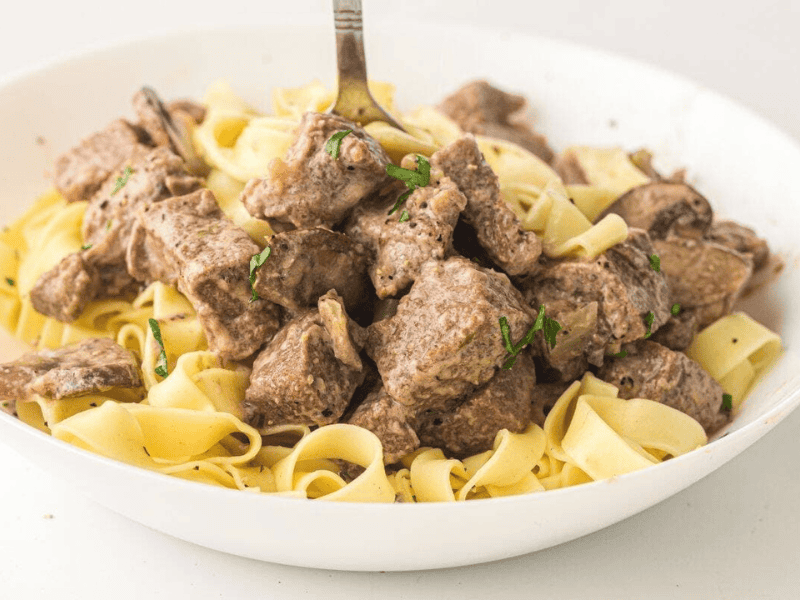 A white bowl filled with cooked egg noodles and chunks of beef, garnished with herbs, with a fork standing upright in the center.