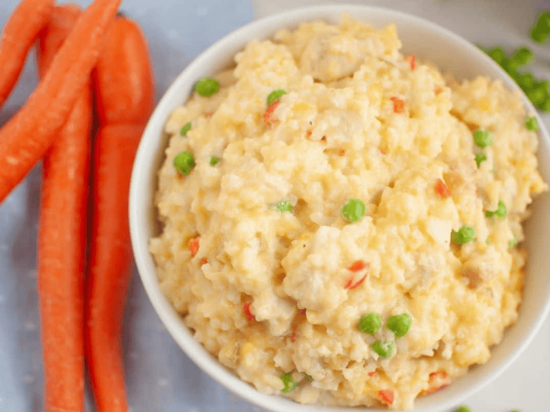 A bowl of Slow Cooker Chicken and Rice is next to whole carrots on a light blue cloth.