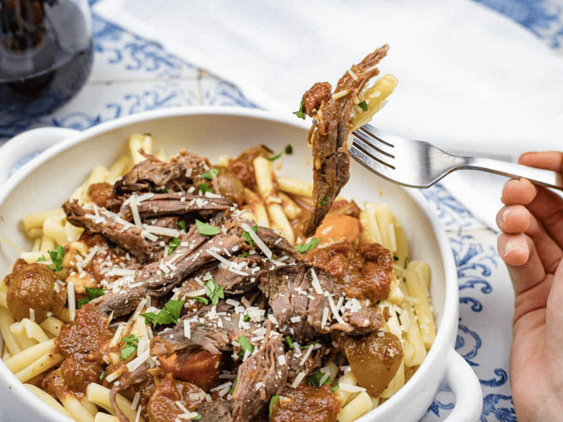 A hand holds a fork with Italian Pot Roast Pasta in a white bowl, topped with grated cheese and herbs.