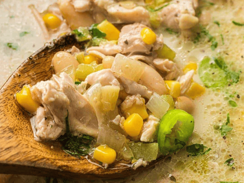 A wooden spoon holding a portion of white chicken chili with corn, beans, onions, and herbs.