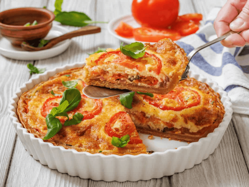 A slice of tomato pie being lifted with a serving utensil from a white fluted dish. A whole tomato pie is visible in the dish, garnished with tomato slices and basil.