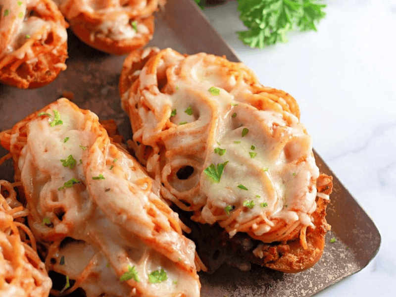 Toasted bread slices topped with spaghetti, meat sauce, melted cheese, and garnished with chopped parsley on a metal baking sheet.