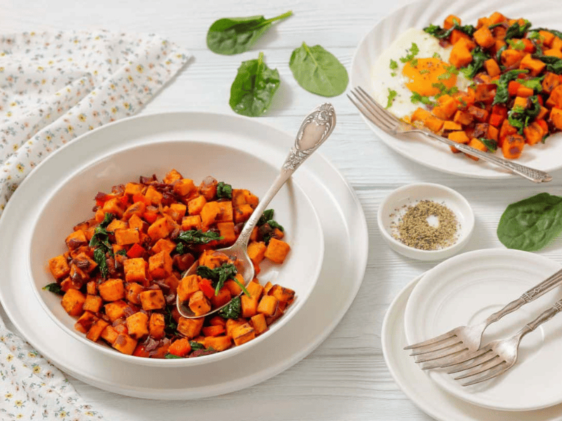 A bowl and plates of sweet potato hash with saut&eacute;ed spinach and onions, served on a white table with forks, a napkin, and a small bowl of pepper.
