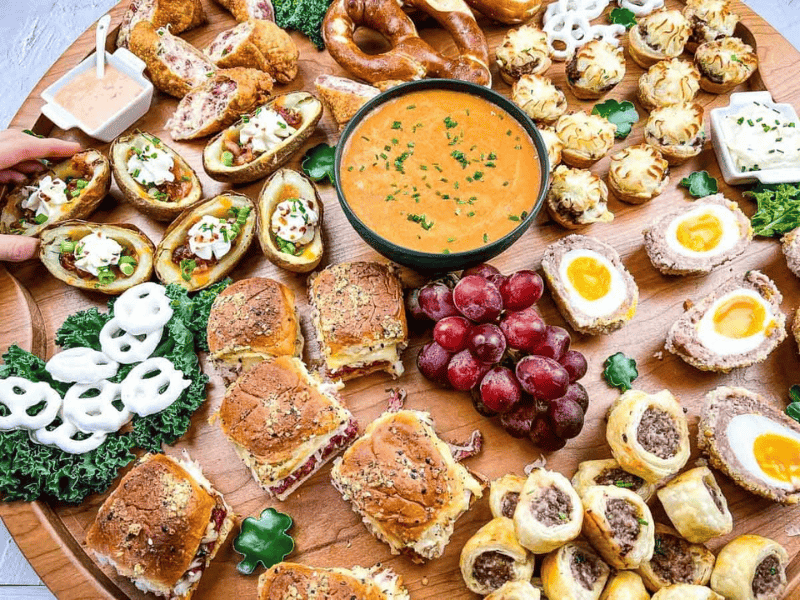 A round wooden platter filled with various appetizers including sliders, stuffed potato skins, Scotch eggs, pretzels, grapes, and dipping sauce.