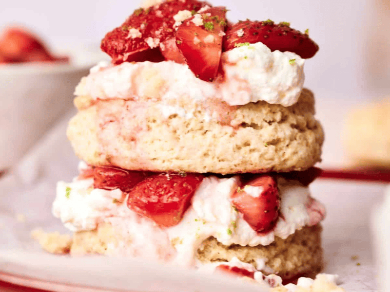 A close-up of a delectable strawberry shortcake reveals layers of biscuit, whipped cream, and fresh strawberry pieces on a plate. In the background, a bowl of strawberries enhances the sweet temptation.