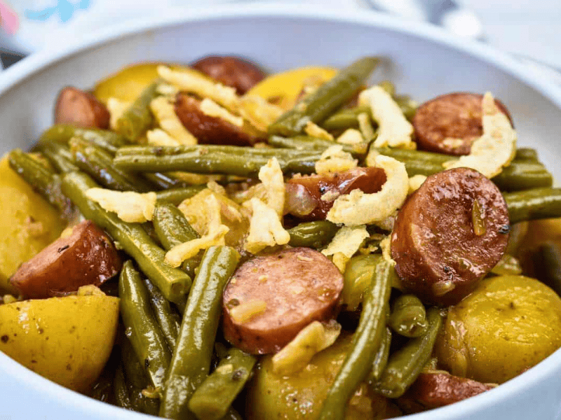 A bowl of Swamp Potatoes with cooked green beans, sliced sausage, and halved potatoes, topped with crispy fried onions.