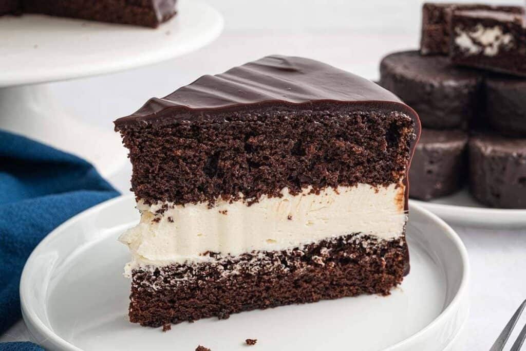 A slice of chocolate cake with a thick layer of cream filling sits on a white plate, surrounded by even more tempting chocolate treats in the background.
