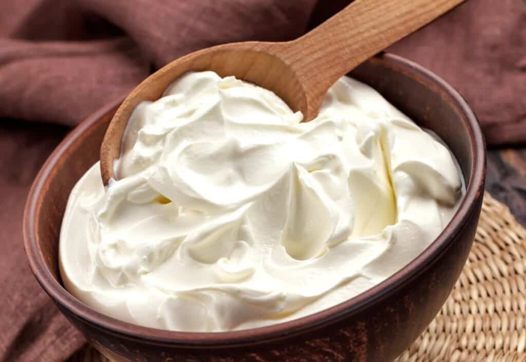 A wooden spoon rests in a brown bowl filled with thick, creamy white yogurt on a woven placemat with a brown cloth in the background.