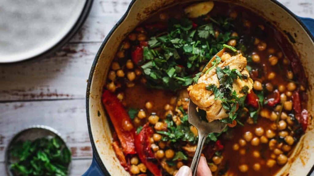 A close-up of a pot with chicken stew, chickpeas, red peppers, and fresh herbs, with a fork lifting a portion from the dish.