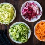 Four bowls containing spiralized vegetables—zucchini, beet, cucumber, and carrot—are arranged on a wooden table next to a black checkered cloth.