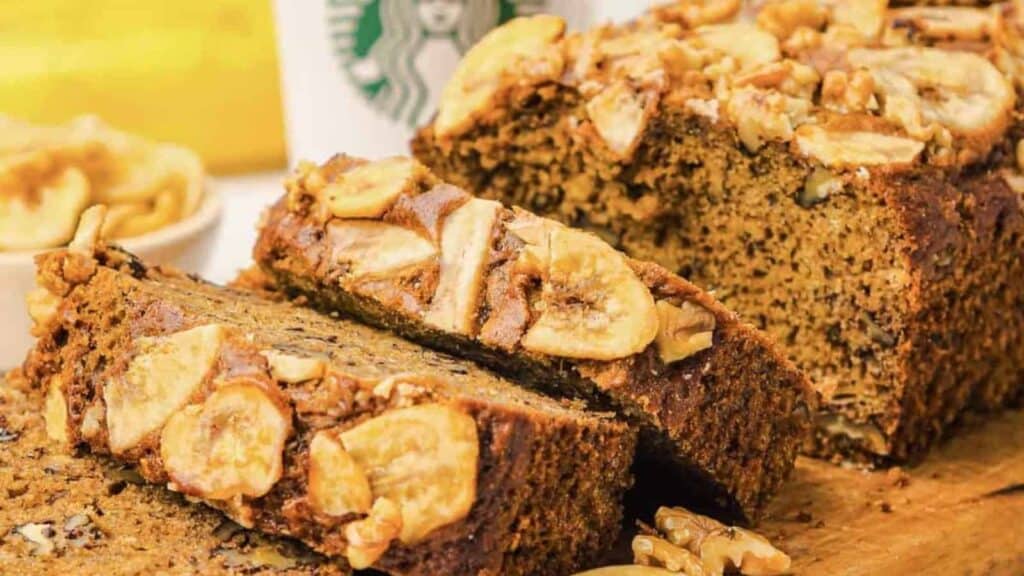 A sliced loaf of banana nut bread with walnuts and banana chips on top sits on a wooden board; a Starbucks cup is in the background.