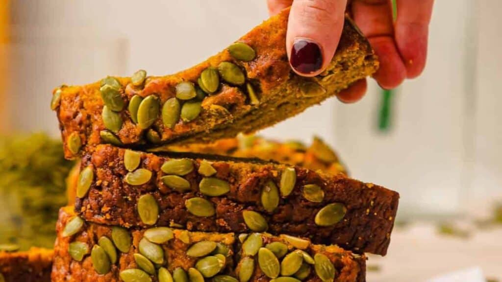 A hand picking up a slice of bread topped with pumpkin seeds from a stack of similar slices.