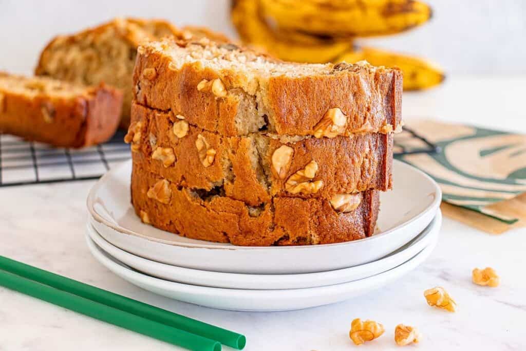Three slices of banana nut bread are stacked on a white plate, with bananas and another loaf in the background and green straws nearby&mdash;perfect for fans of Starbucks Copy Cat Recipes.
