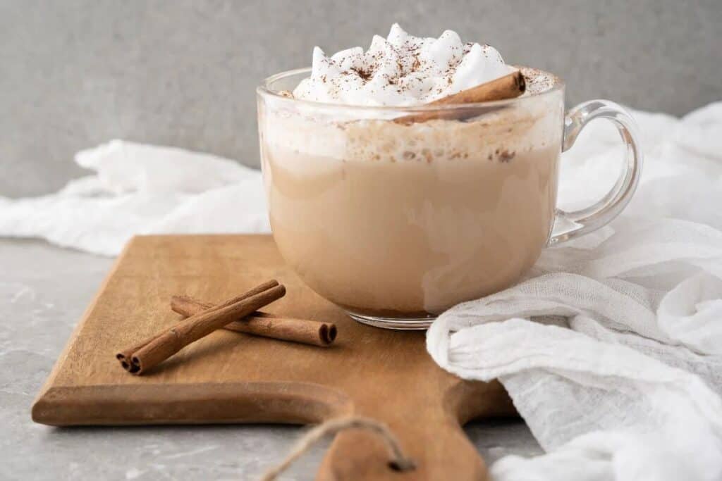 A glass mug of creamy latte topped with whipped cream and a dusting of cinnamon, garnished with a cinnamon stick, sits on a wooden board beside two loose cinnamon sticks&mdash;perfect for Starbucks Copy Cat Recipes lovers.