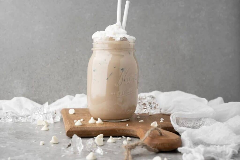 A mason jar filled with iced coffee topped with whipped cream and two straws, placed on a wooden board with scattered white chocolate chips&mdash;perfect for trying your favorite Starbucks Copy Cat Recipes at home.