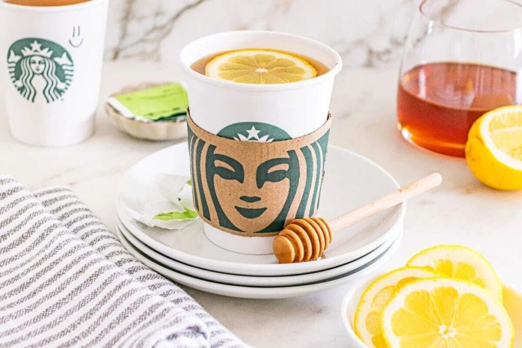 A Starbucks cup with a lemon slice in hot tea sits on plates, next to a honey dipper, lemon slices, a tea bag, and a carafe of tea&mdash;perfect for recreating your favorite Starbucks Copy Cat Recipes at home.