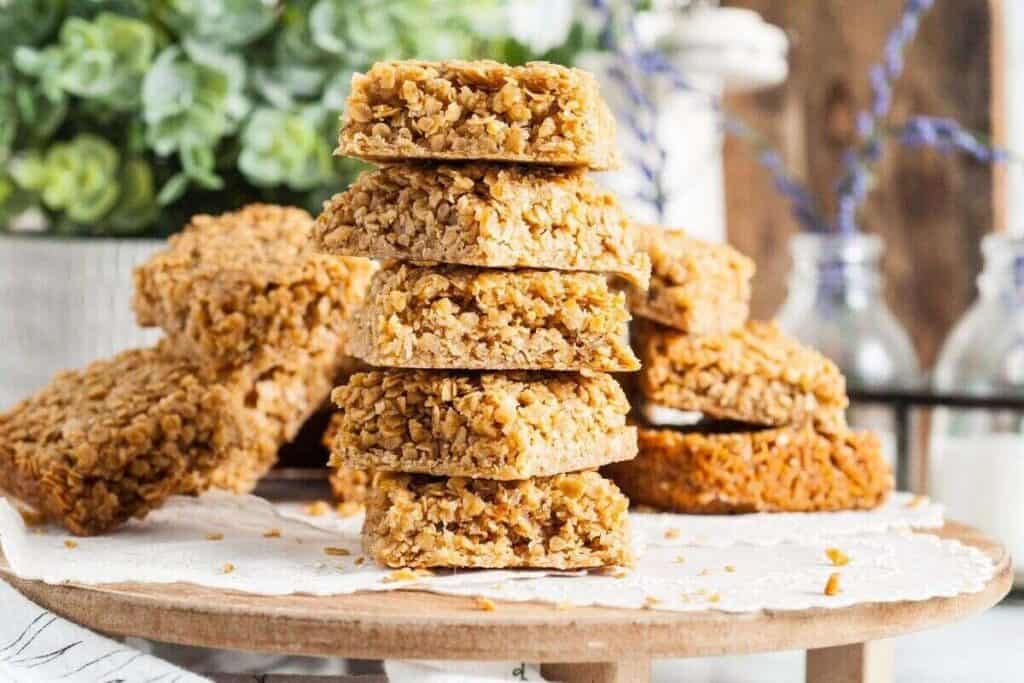 A stack of oat flapjacks sits on a wooden platter, surrounded by more flapjacks, perfect for those seeking Starbucks Copy Cat Recipes, with blurred plants and bottles in the background.