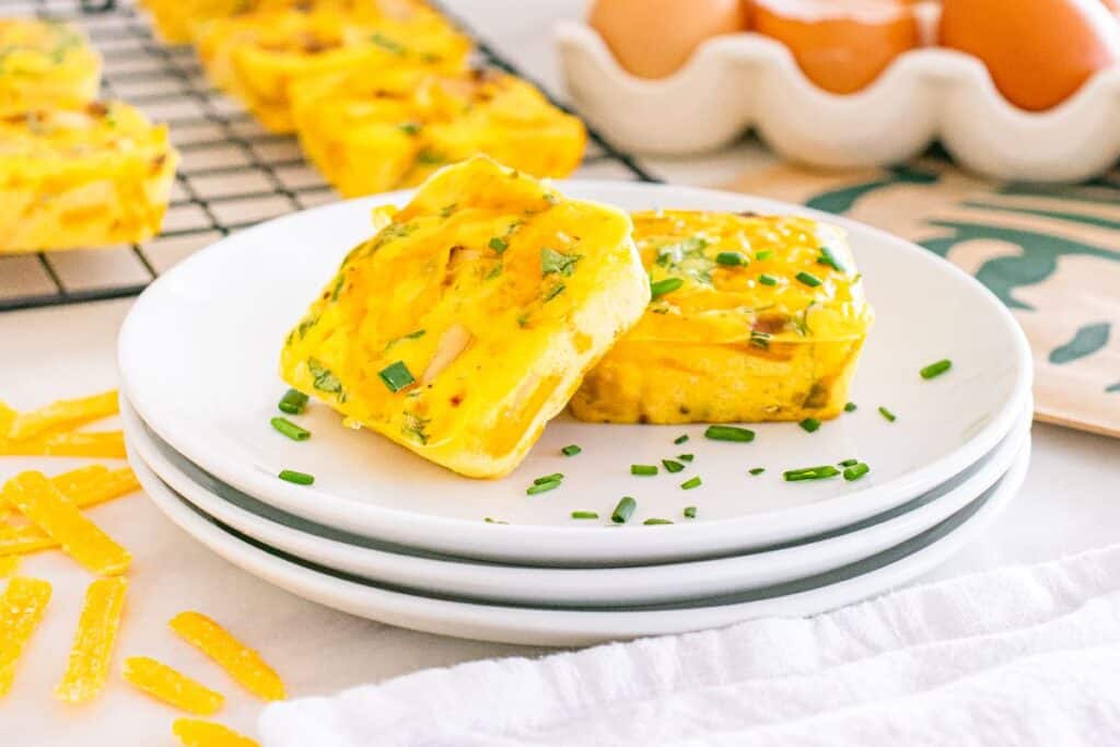 Two square egg muffins garnished with chives are on a white plate. Inspired by Starbucks Copy Cat Recipes, more muffins, eggs in a carton, and shredded cheese can be seen in the background.