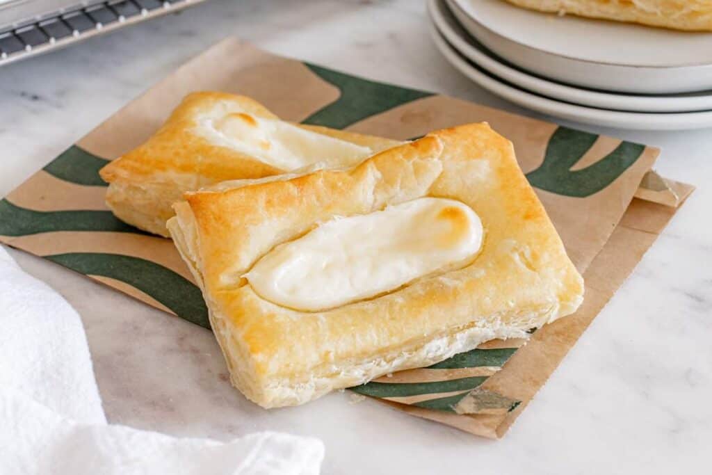 Two rectangular pieces of puff pastry with a creamy white filling in the center, placed on a patterned piece of paper&mdash;these treats are perfect for fans of Starbucks copy cat recipes.
