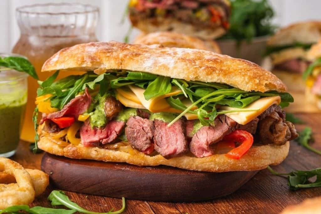 A sandwich with sliced steak, cheddar cheese, arugula, red bell peppers, and sauce on ciabatta bread&mdash;one of our tasty beef recipes&mdash;served on a wooden board.