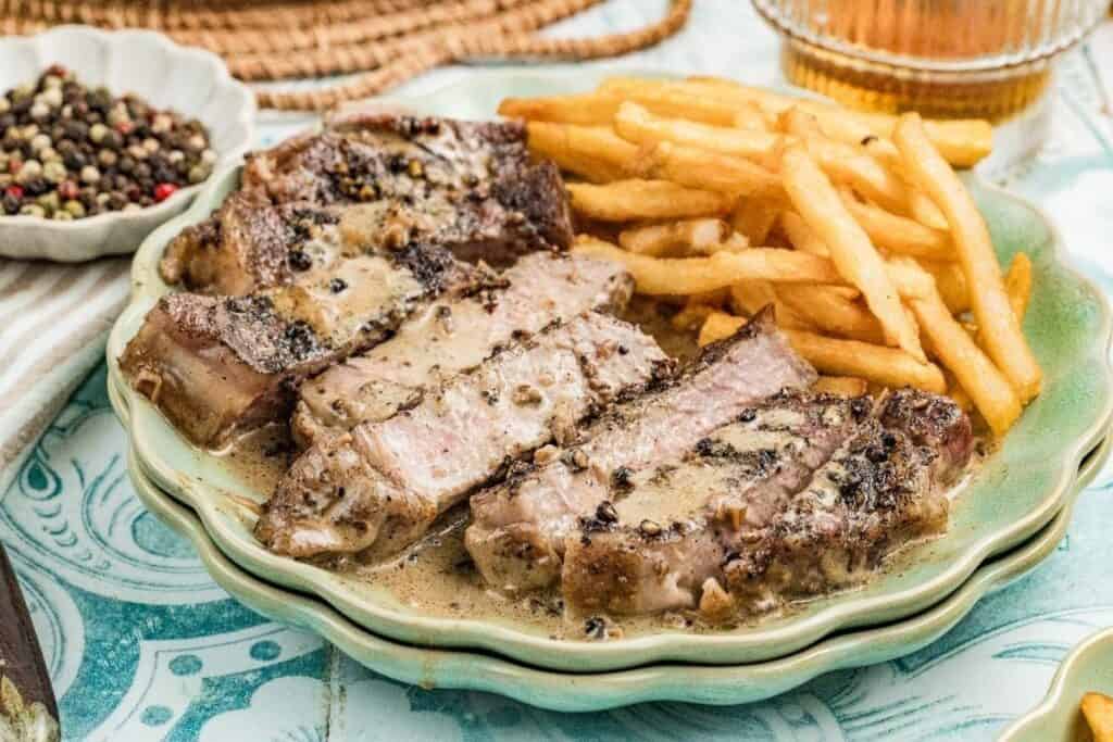 A plate of sliced steak with creamy peppercorn sauce, served alongside French fries&mdash;one of the tastiest beef recipes to try. A bowl of mixed peppercorns and a drink are visible in the background.