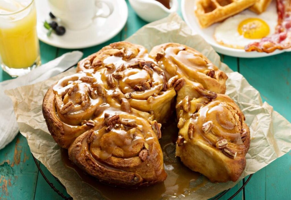 A batch of pecan sticky buns with caramel glaze on parchment paper, perfect for celebrating National Sticky Bun Day, with a breakfast plate, cup, and glass of orange juice in the background.