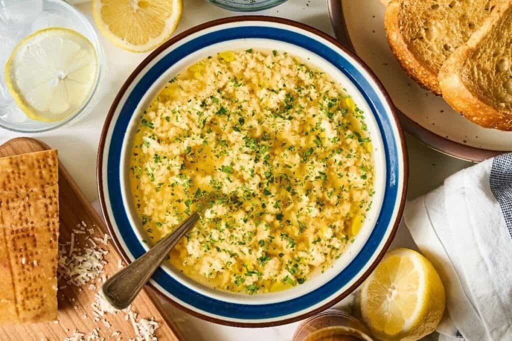 A bowl of hummus topped with chopped herbs and olive oil, surrounded by lemon halves, toasted bread, and a glass of water with lemon slices&mdash;perfect for easy recipes made for busy schedules.