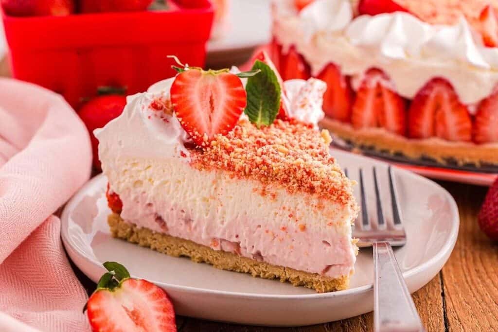 A slice of strawberry cream pie topped with whipped cream, crushed crumbs, and a fresh strawberry, served on a white plate with a fork&mdash;perfect for fans of simple cheesecake desserts.