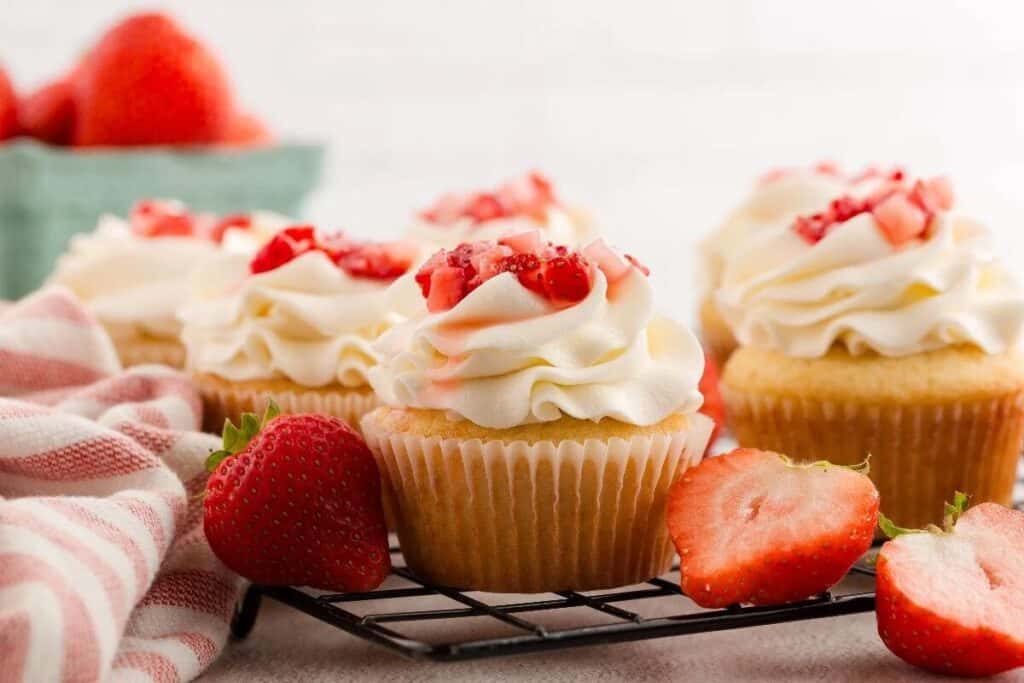 Vanilla cupcakes topped with whipped cream and chopped strawberries, displayed on a cooling rack with whole and halved strawberries nearby.