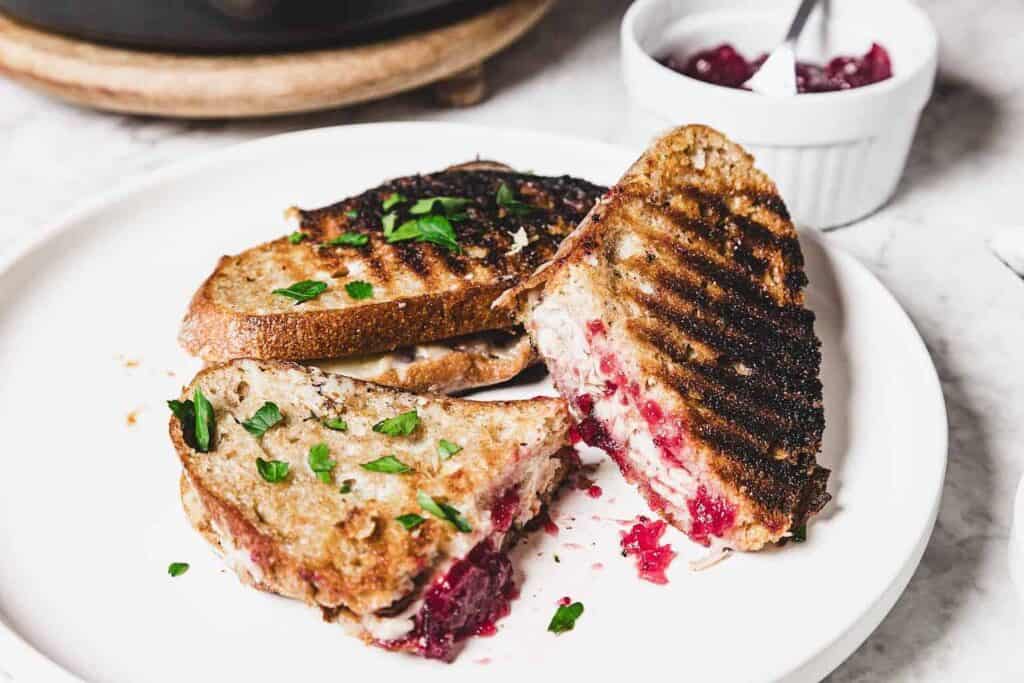 A grilled sandwich with melted cheese and cranberry filling is cut in half on a white plate, garnished with chopped herbs. A small bowl of cranberry sauce is in the background.
