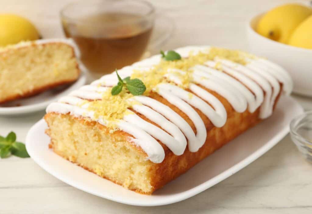 A lemon loaf cake with white icing and lemon zest is served on a white plate, with a cup of tea, lemons, and a slice of cake in the background.