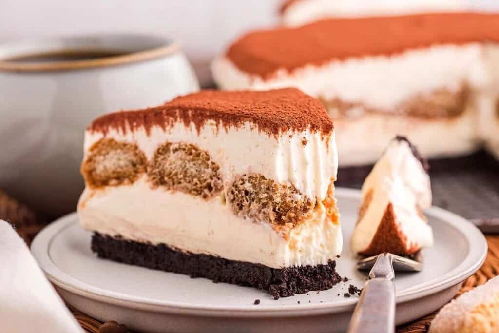 A slice of layered tiramisu simple cheesecake with a dusting of cocoa powder sits on a plate with a fork. A cup and the whole cheesecake are in the background.