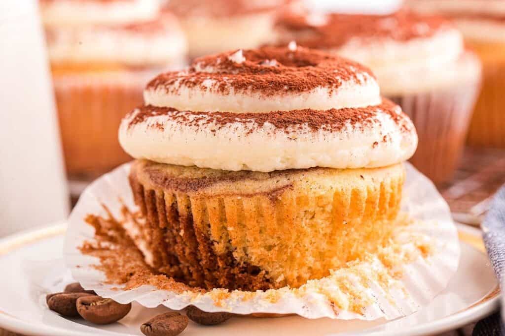 A cupcake with white frosting and cocoa powder sits on a plate, partially unwrapped with a bite taken out. Coffee beans are scattered nearby.