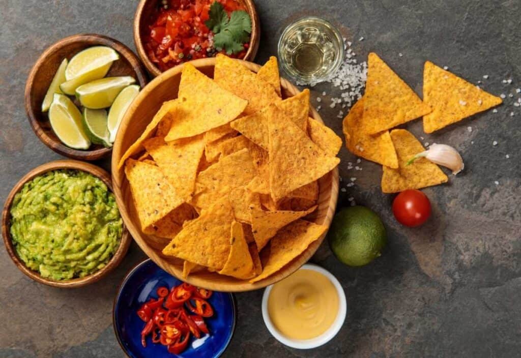 Bowl of tortilla chips surrounded by guacamole, salsa, lime wedges, sliced chili, cheese dip, garlic, a whole lime, a cherry tomato, and a glass of clear beverage on a dark surface.