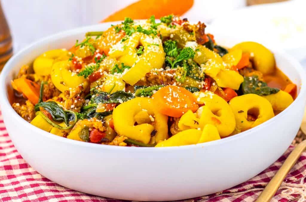 A bowl of tortellini soup with vegetables and herbs, topped with grated cheese, sits on a red checkered cloth.