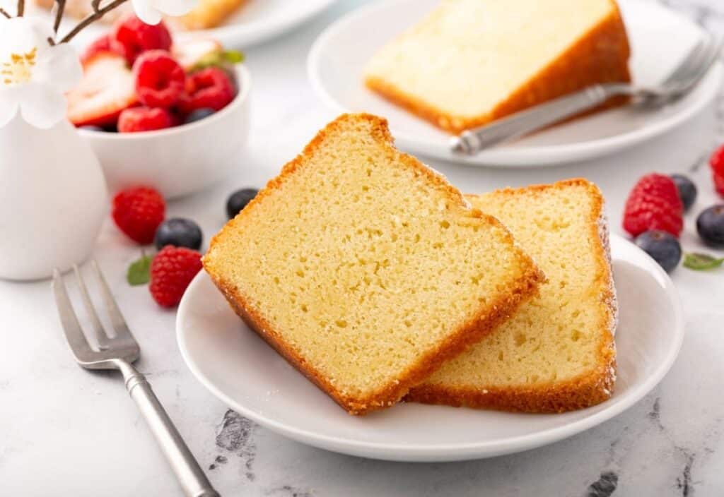 Two slices of plain pound cake on a white plate, with a fork beside them and a bowl of mixed berries in the background—perfect for celebrating National Pound Cake Day.