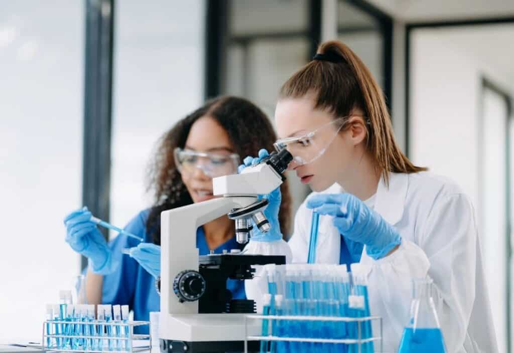 Two women wearing lab coats, gloves, and safety goggles work in a laboratory; one looks into a microscope while the other handles a pipette near test tubes.
