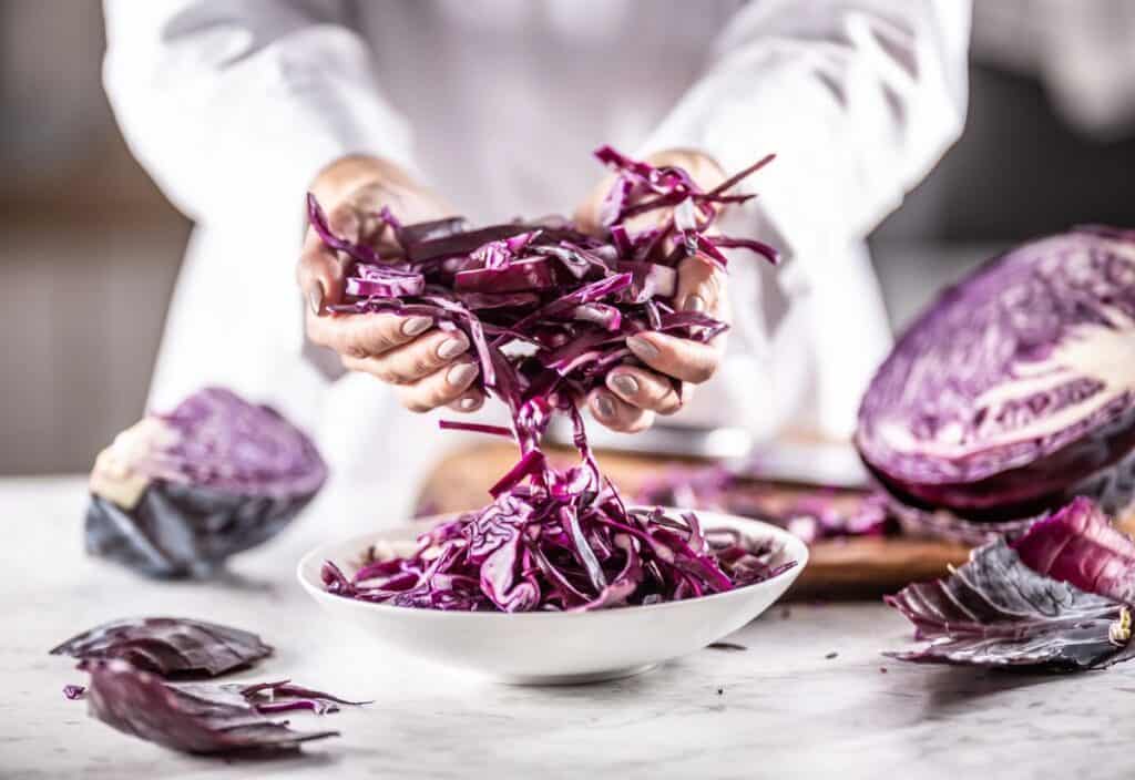 A person places shredded red cabbage into a white bowl, with more cabbage and a knife visible on a cutting board in the background.
