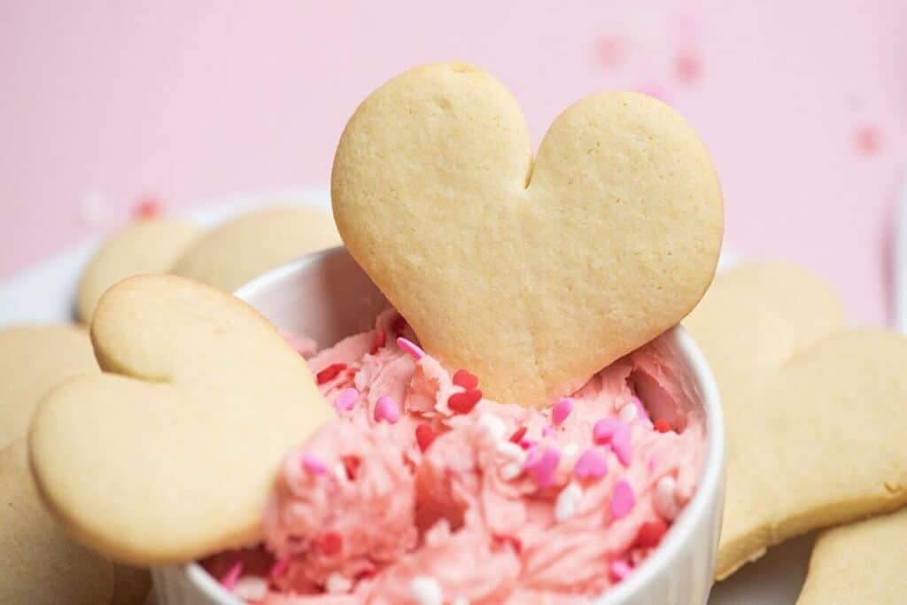Heart-shaped sugar cookies with a bowl of pink frosting and heart-shaped sprinkles make delightful Valentines Treats on a light pink background.