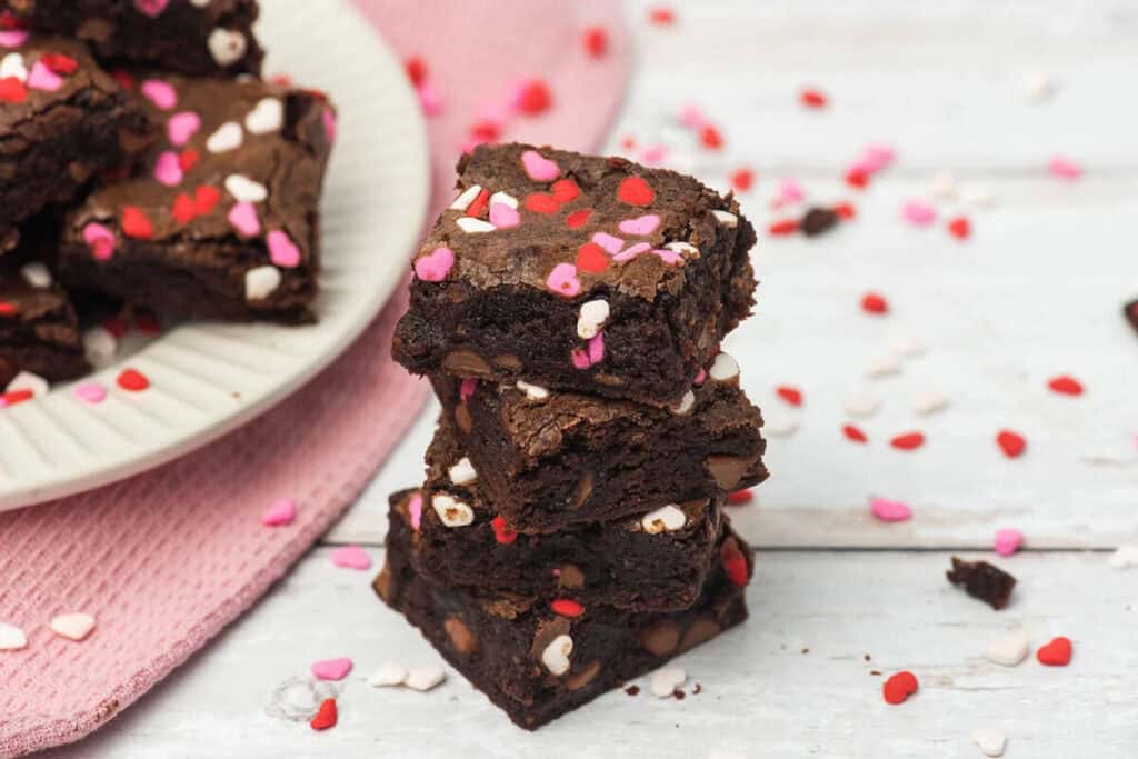A stack of chocolate brownies topped with pink, red, and white heart-shaped sprinkles sits on a white wooden surface beside a plate of more Valentine’s treats and a pink cloth.