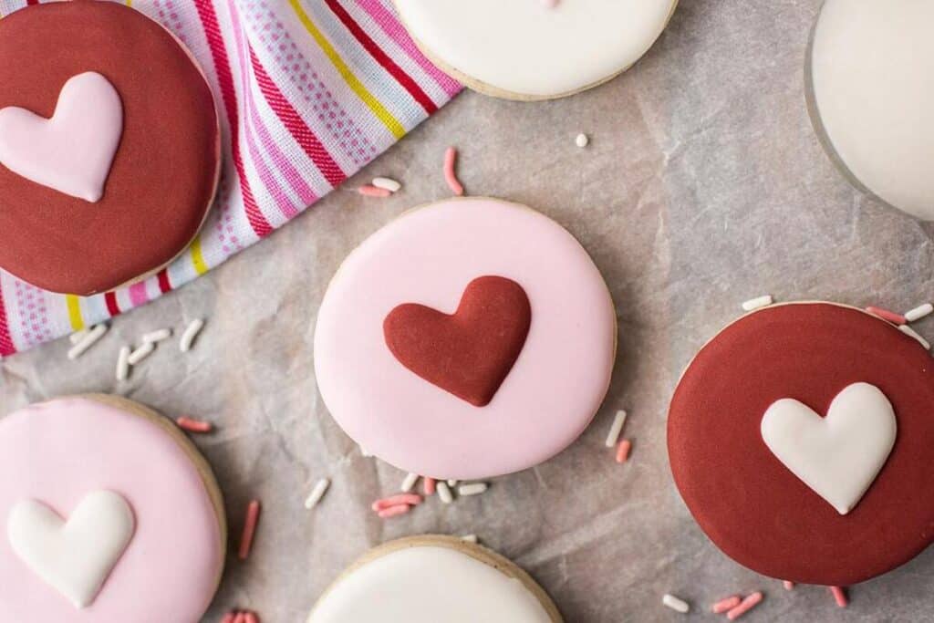 Round cookies decorated with red and white fondant hearts are arranged on parchment paper next to a striped cloth, with scattered sprinkles around them—perfect Valetines Treats for sharing.