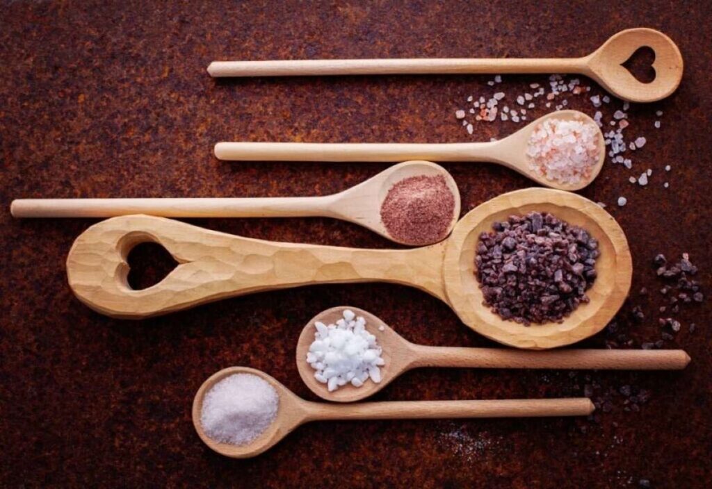 Six wooden spoons with heart-shaped cutouts, each holding a unique salt or spice, are artfully arranged on a dark brown surface.