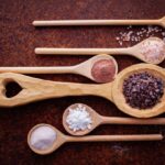 Six wooden spoons with heart-shaped cutouts, each holding a unique salt or spice, are artfully arranged on a dark brown surface.