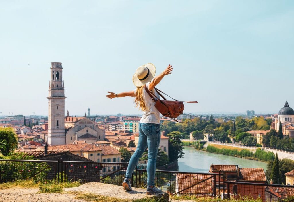 A woman with a hat and backpack stands on a hill with arms outstretched, overlooking a cityscape with historic buildings and a river.