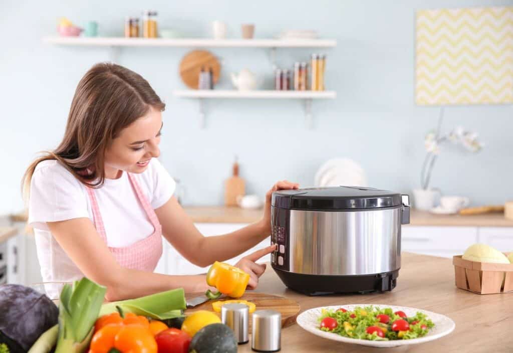 Woman in a kitchen pressing buttons on a stainless steel multi-cooker, surrounded by fresh vegetables and a prepared salad—perfect for quick, lazy dinners.