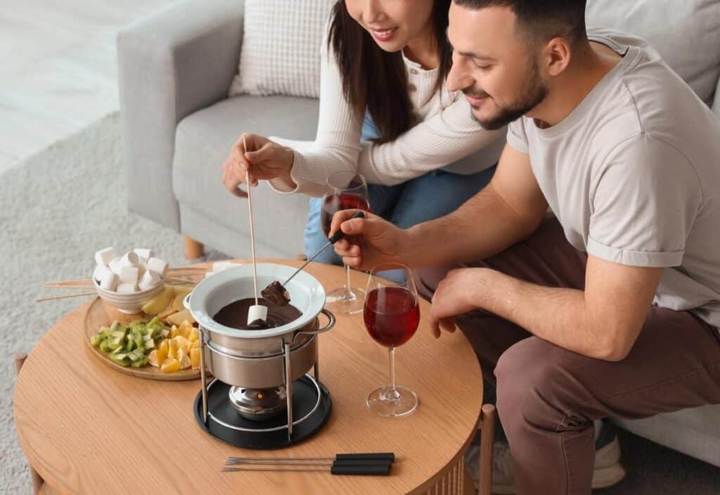 Two people sit at a coffee table dipping marshmallows into a chocolate fondue pot, with fruit and glasses of red wine nearby.