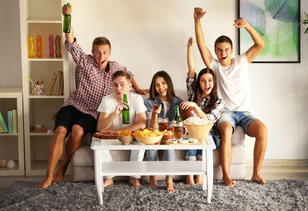 Five young adults sit on a couch watching TV, cheering and holding drinks, with snacks like popcorn and chips on a table in front of them.